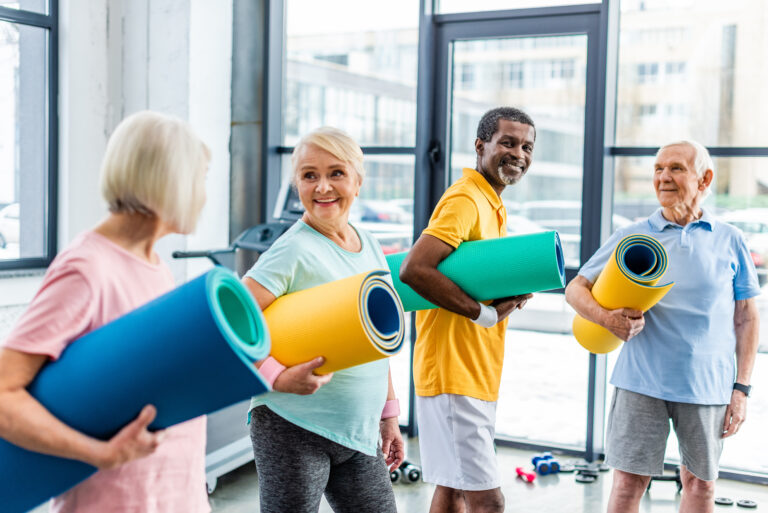 happy multethnic senior athletes holding fitness mats at sports hall